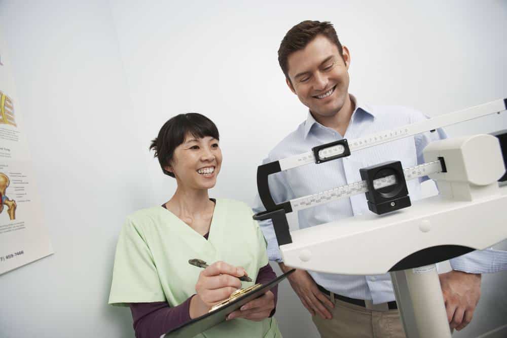 Man Measuring Weight With Doctor Noting On Clipboard