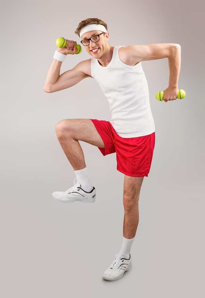 Geeky hipster posing in sportswear with dumbbells on grey background