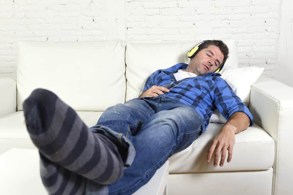 man falling asleep on home couch while listening to music with mobile phone and headphones